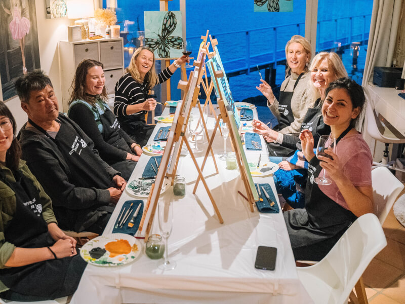 A group of six women and one man wearing aprons sit smiling at a table in an art studio overlooking some water. They each have an easel, glasses of drinks and painting materials.
